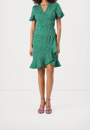 Femme portant une robe portefeuille verte à manches courtes avec un motif floral blanc et un ourlet à volants, associée à des sandales à talons violet à fines brides.