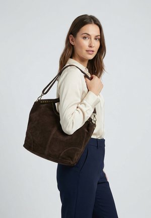 Frau trägt cremefarbene Bluse und marineblaue Hose, trägt eine große, dunkelbraune Wildleder-Schultertasche mit Messingnieten.