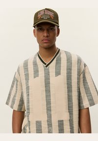 Beige and gray striped knit short-sleeve shirt with a V-neck and a large beige "11" patch on the front, worn with an olive green cap.