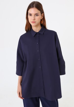 Giovane donna che indossa una camicia blu navy larga con bottoni, maniche a tre quarti e orecchini rotondi marroni su uno sfondo semplice.