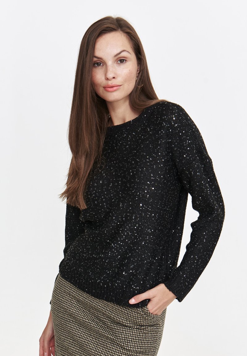 Black sweater with metallic shimmer, round neck, long sleeves, and a textured pattern. Worn with a patterned skirt, showcasing casual layering.