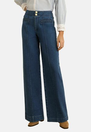 Weit geschnittene dunkelblaue Jeans mit geflochtenem Taschen-Detail, hoher Taille mit doppelten goldenen Knöpfen, kombiniert mit einer gestreiften Bluse und braunen spitzen Schuhen.