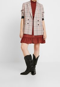 Blazer de cuadros de doble botonadura en gris, blanco y rojo; vestido burdeos en capas con dobladillo volado; botas de vaquero de cuero negras con punta afilada.