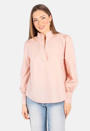 Smilende kvinne med rett brunt hår har på seg lys rosa bluse med lange ermer og lyseblå jeans mot hvit bakgrunn.