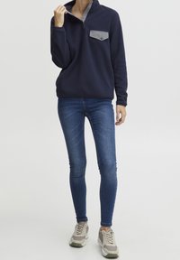 Sudadera de forro polar azul marino con cuello de media cremallera y bolsillo en el pecho gris, combinada con jeans ajustados azules y zapatillas de deporte gris claro.