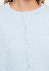 Ανοιχτό μπλε πουλόβερ φτιαγμένο από μαλακό ύφασμα, με στρογγυλή λαιμόκοψη, έξι κουμπιά μπροστά και ομαλή υφή με διακριτό σχέδιο.