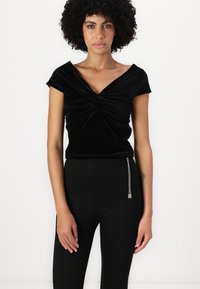 Top de terciopelo negro con un diseño de nudo retorcido en el frente, con mangas cortas y un escote amplio. Combinado con pantalones negros ajustados.