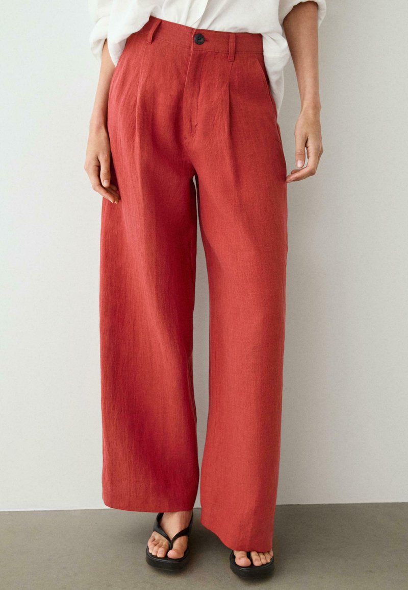 Vrouw draagt rode linnen broek met hoge taille en wijde pijpen met voorplooien, gecombineerd met zwarte slippers en een ingestoken wit overhemd.
