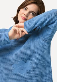 Helles blaues Strickoberteil mit lockerer Passform. Das Oberteil weist ein strukturiertes Muster und ein Blumendetail auf der Vorderseite auf.