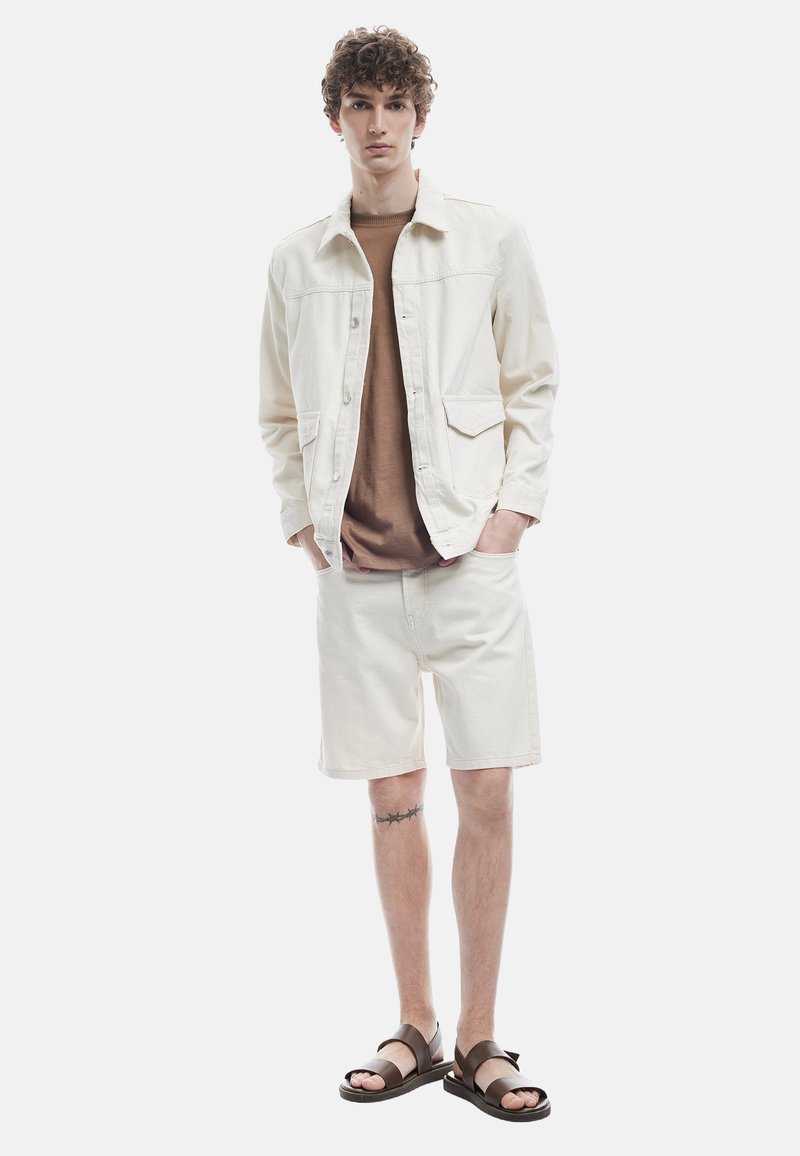 Chaqueta de mezclilla de color claro sobre una camiseta marrón, combinada con pantalones cortos claros y sandalias marrones. Presenta bolsillos y un ajuste casual.