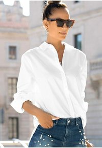 Hvid krave skjorte med knapper, afslappet pasform, parret med slidte blå denimshorts med perleudsmykning og en hånd i lommen.
