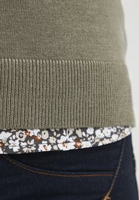 Olivengrøn strikket sweaterkant lagdelt over en blomstret skjorte i dæmpede farver, sat mod mørke denim. Har ribbet tekstur.