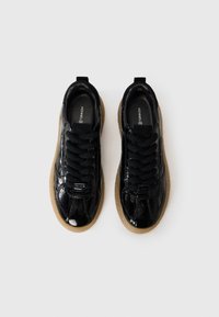 Une paire de baskets noires brillantes avec des semelles en caoutchouc épaisses et des lacets noirs, vues de dessus sur un fond blanc.