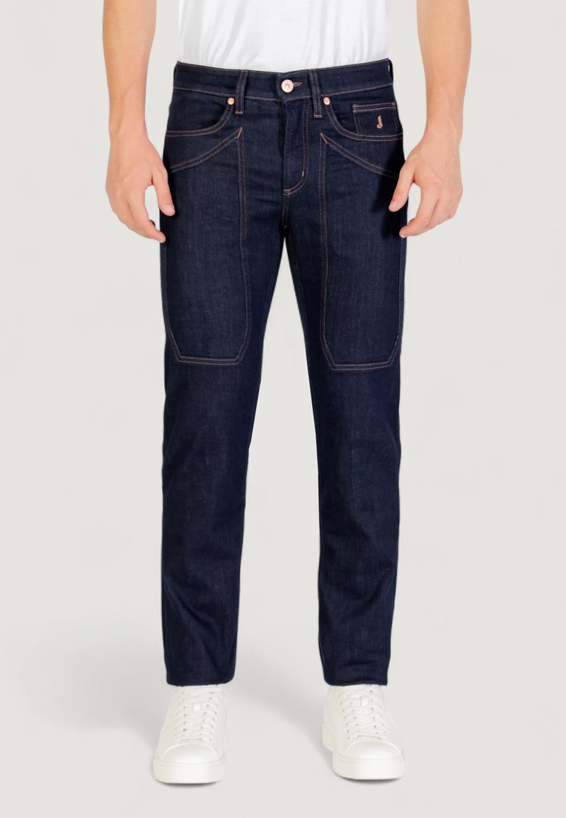 Jean en denim bleu foncé avec des coutures claires, poches avant et coupe droite. Porté avec des baskets blanches et une chemise blanche unie.