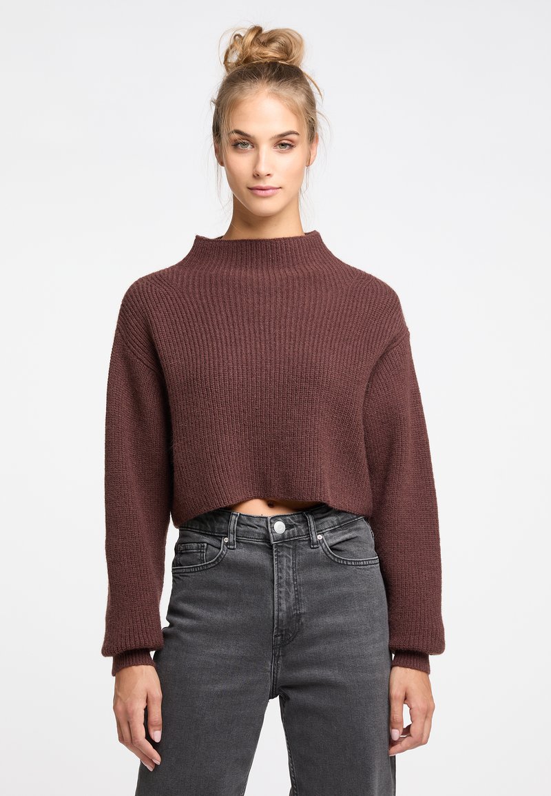 Brauner gerippter Crop-Pullover mit hohem Kragen und langen Ärmeln, kombiniert mit hochgeschnittenen, dunkelgrauen Jeans. Die Textur ist sichtbar und gemütlich.