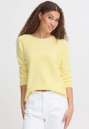 Femme aux cheveux bouclés portant un pull jaune pâle à manches longues et un pantalon blanc, debout devant un fond clair uni.