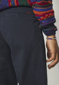 Personne portant un pantalon bleu marine foncé avec une petite étiquette orange "BOSS", un pull coloré en tricot, et un bracelet en argent avec des perles bleues au poignet.