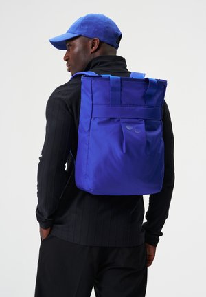 Mann mit einem leuchtend blauen Rucksack und passender Kappe, gekleidet in ein schwarzes Langarmshirt und schwarze Hose, seitlich stehend.