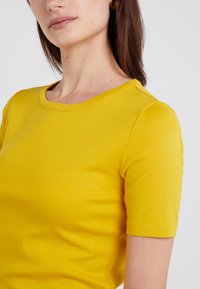Žluté bavlněné tričko s přiléhavým střihem, kulatým výstřihem a krátkými rukávy. Hladká textura bez viditelných vzorů nebo ozdob.