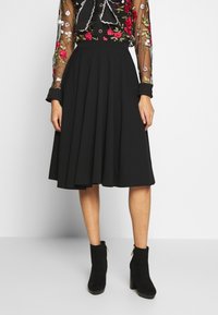 Donna che indossa una gonna midi nera, una blusa nera trasparente con ricami floreali colorati e stivaletti neri in suede con tacco a blocco.