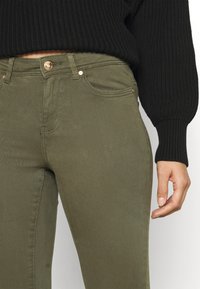 Jeans verde-oliva feitos de um tecido de algodão misto, com um design de cinco bolsos, fecho de botão de metal e costuras detalhadas ao longo das costuras.