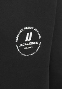 Crna majica s okruglim bijelim logom koji sadrži "JACK&JONES" i "RESEARCH_DESIGN_APPAREL." Glatka tkanina s prilagođenim dizajnom.