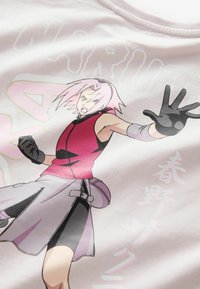 Rožinė medvilninė marškinė, vaizduojanti veikėją su rožiniais plaukais, apsirengusį raudona liemene, juodomis pirštinėmis ir pilka sijonu, su japonų kalbos užrašu.