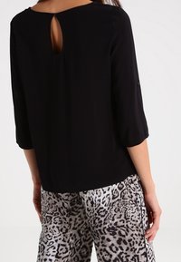 Schwarze Bluse mit drei Viertelärmeln, mit einem runden Halsausschnitt und einem Schlüssellochdetail im Rücken. Kombiniert mit gemusterten Leopardenprint-Hosen.