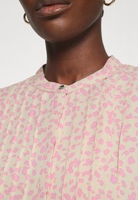 Bluzka w jasnobeżowym materiale z różowym wzorem w panterkę, z kołnierzykiem mandarynkowym i zapięciem na guziki przy dekolcie. Widoczne teksturowane fałdy.