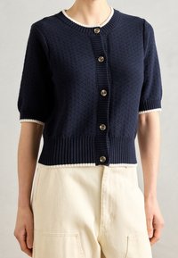 Navy knitted cardigan with short sleeves, ribbed hem and cuffs, five button closure, and white trim around the neckline and cuffs.