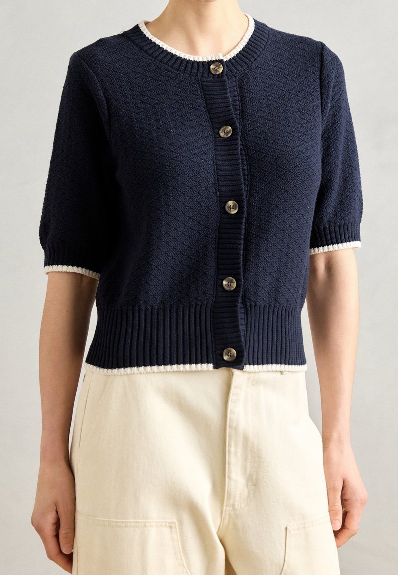Navy knitted cardigan with short sleeves, ribbed hem and cuffs, five button closure, and white trim around the neckline and cuffs.
