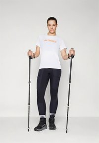 Biała koszulka Terrex z pomarańczowym logo, czarne dopasowane legginsy z kieszeniami oraz czarne buty trekkingowe. Użytkownik trzyma kije trekkingowe.