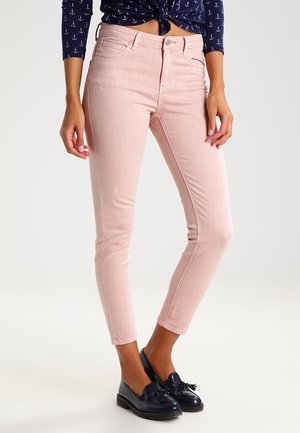 Frau trägt hellrosa, figurbetonte Jeans, marineblaues, mit Anker-Muster versehenes gebundenes Hemd und schwarze Loafers mit Quasten, steht vor einem einfarbigen Hintergrund.