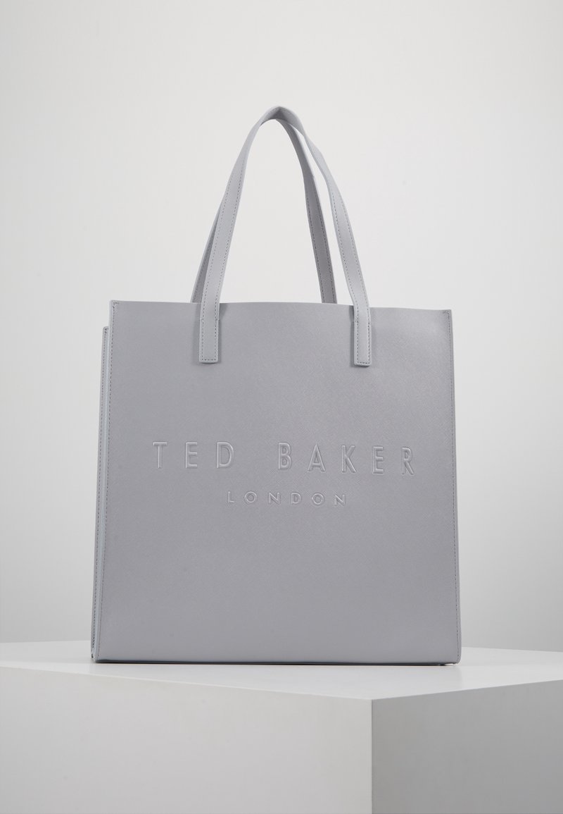 Sac fourre-tout gris Ted Baker London avec deux poignées, debout sur un piédestal blanc sur un fond gris uni.