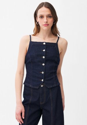 Jeune femme portant un haut caraco en jean bleu foncé à boutons avec des bretelles fines et un pantalon en jean assorti taille haute.