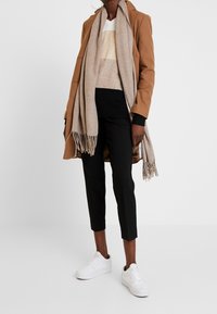 Cappotto marrone, sciarpa beige, maglione a righe multicolori, pantaloni neri e sneakers bianche. Tessuti morbidi, design a strati e dettagli frangiati sulla sciarpa.