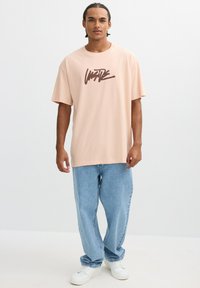 Camiseta oversized de color rosa claro con letras gráficas marrones, combinada con unos pantalones de mezclilla azules holgados y zapatillas blancas. Diseño minimalista y ajuste casual.