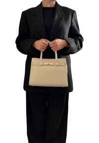 Osoba w czarnym garniturze trzyma beżową skórzaną torebk�ę z złotym zapięciem na tle białym.