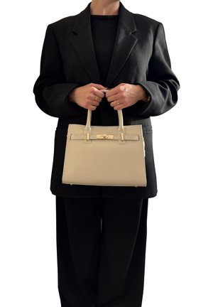 Persona che indossa un abito nero e tiene una borsa in pelle beige con chiusura dorata davanti a uno sfondo bianco.