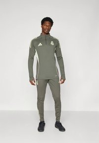 Chándal deportivo verde oliva con camiseta de cremallera de cuarto y pantalones con cintura elástica; presenta detalles en amarillo y logo del equipo en el pecho y el muslo.