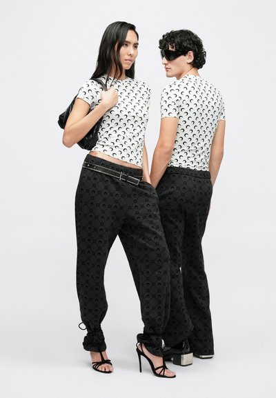 Camisetas blancas de manga corta con patrón repetido de lunas crecientes, combinadas con pantalones jogger negros con un sutil patrón de logotipos, y sandalias de tacón.