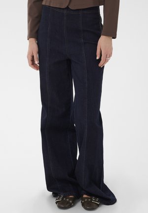 Person iført mørkeblå jeans med vide ben og frontsyninger, brun jakke og sorte flade sko med spænder, stående på hvid baggrund.