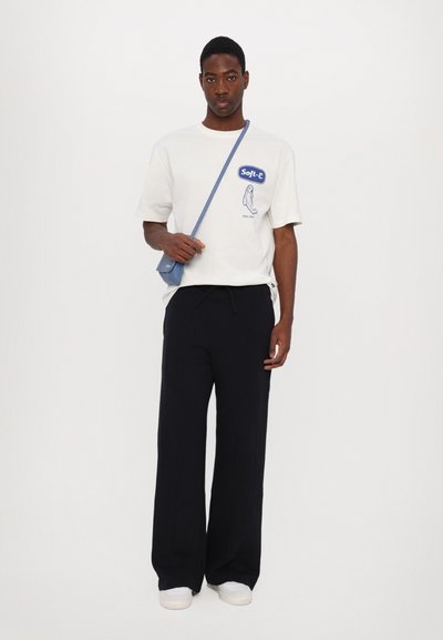 T-shirt en coton blanc avec un imprimé graphique bleu, associé à un pantalon large noir et des baskets blanches, complété par un petit sac bandoulière bleu.
