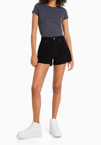 Shorts de mezclilla negros con cintura alta, que cuentan con bolsillos frontales y cierre de botón, combinados con una camiseta ajustada gris oscuro y zapatillas blancas.