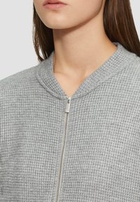 Veste zippée grise texturée avec un col arrondi, dotée d'une fermeture éclair métallique et d'un motif gaufré sur l'ensemble du tissu.