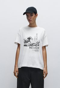 Bílé bavlněné tričko s černou vlnou a japonským textem. Oversized střih s krátkými rukávy. Model má na sobě černou čepici.