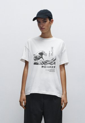 Hvid bomulds t-shirt med et sort bølgemotiv og japansk tekst. Oversized pasform med korte ærmer. Modellen bærer en sort kasket.