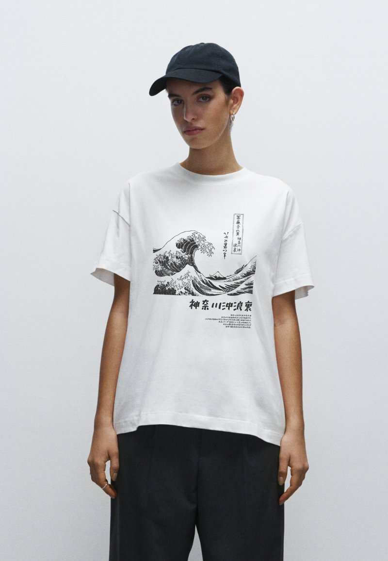 Bílé bavlněné tričko s černou vlnou a japonským textem. Oversized střih s krátkými rukávy. Model má na sobě černou čepici.