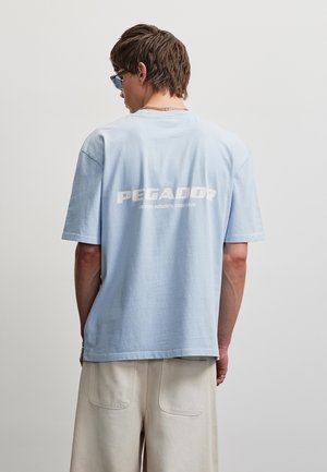 Jeune homme portant un t-shirt bleu clair avec le texte blanc "PEGADOR" dans le dos et un pantalon beige, debout sur un fond gris uni.