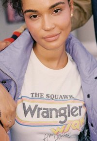 Lila Jacke über einem weißen T-Shirt mit den Logos "The Squawk" und "Wrangler" in Retrofarben und einem charakteristischen Detail.